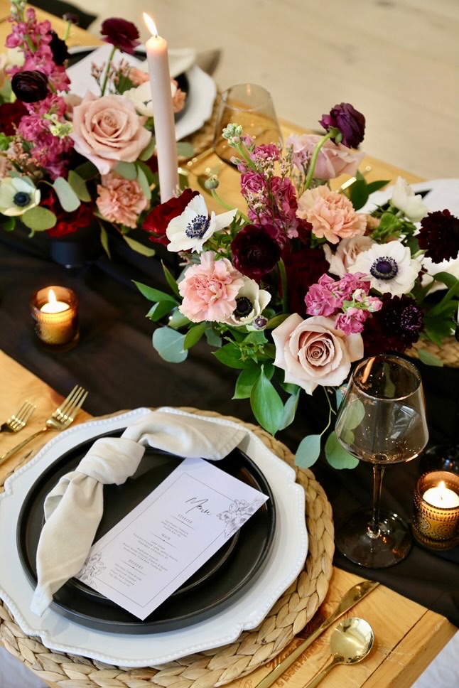 Table centrepiece of blush roses, burgundy dahlias, pink stock and white anemones with candles and a place setting