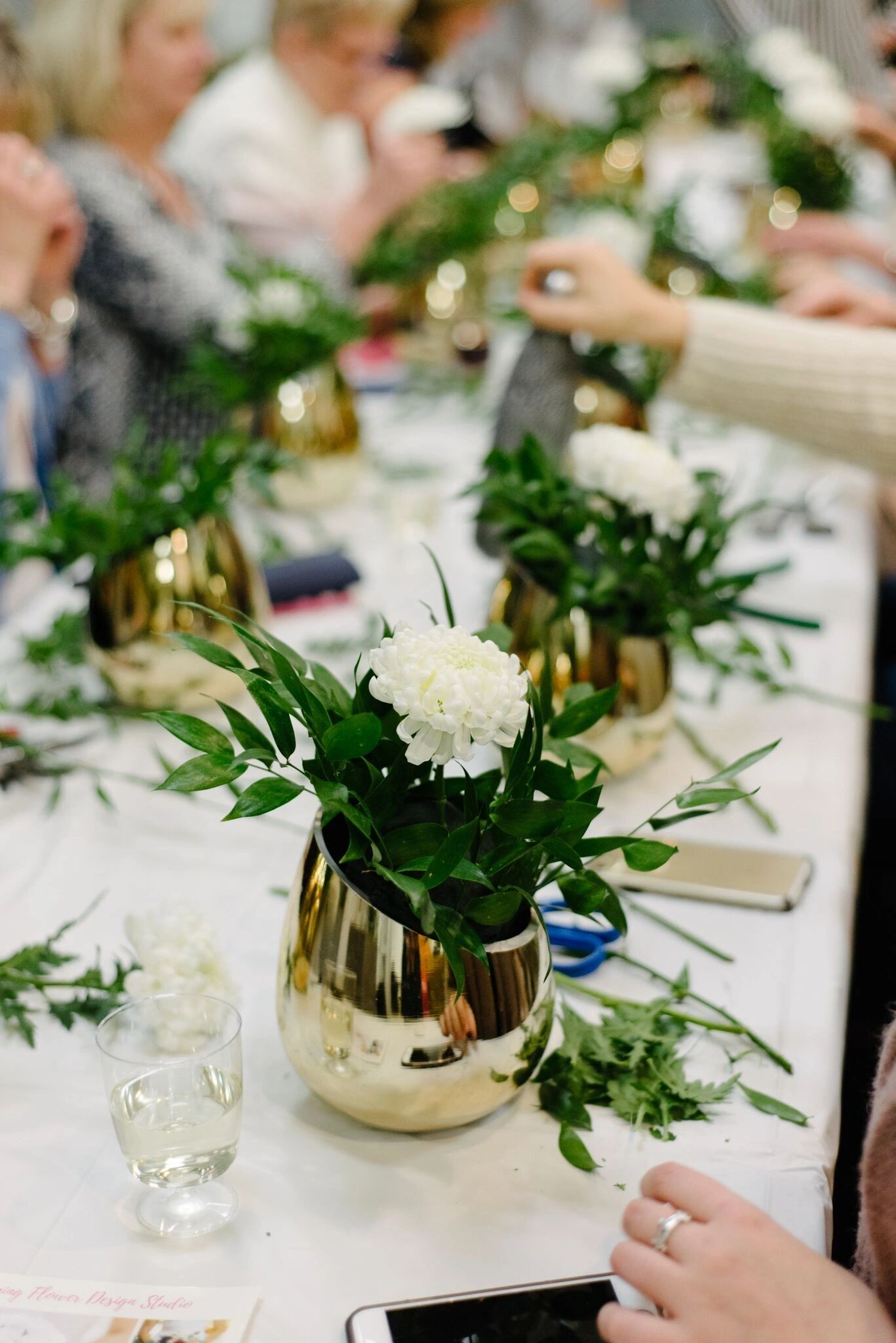 Gold vases of white roses and greenery taking shape along the workshop table