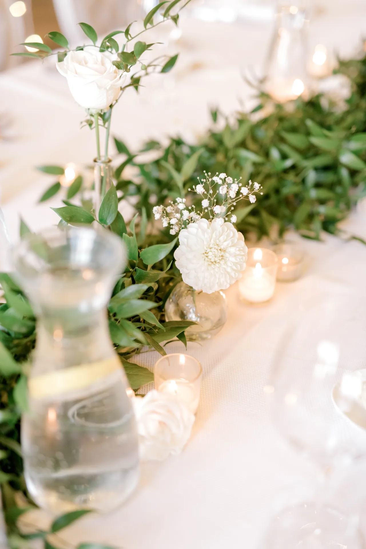 Single white rose and dahlia bud vases with baby’s breath on a greenery garland and candlelight