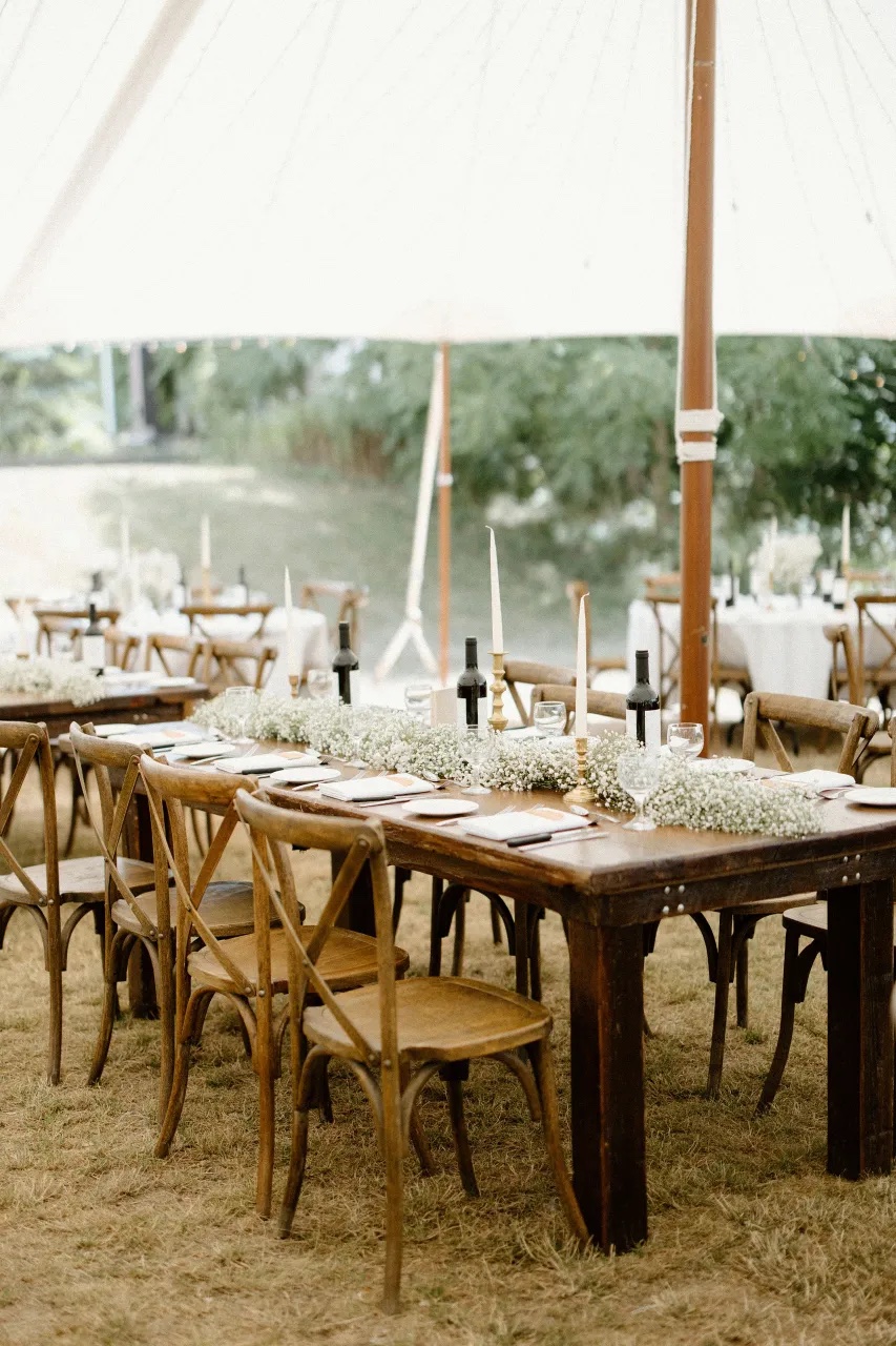 Tented reception with wooden harvest tables, crossback chairs and baby’s breath runners with taper candles
