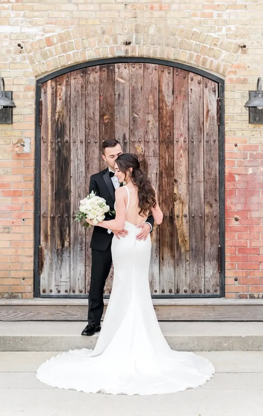 Couple embracing in front of arched wooden doors with a white rose bouquet