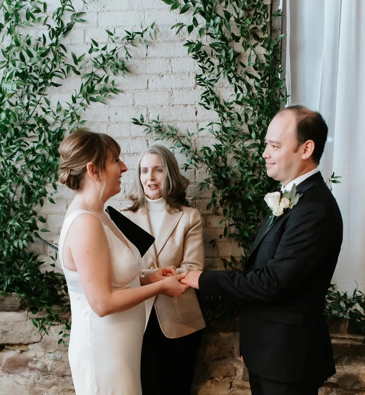 Couple exchanging vows before a white brick wall draped with Italian ruscus