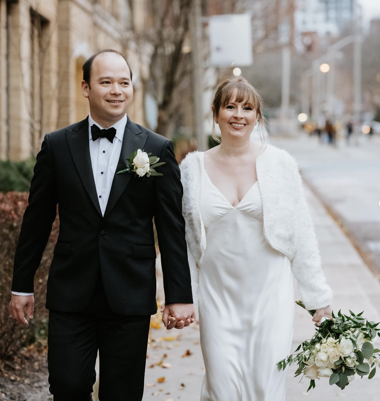 Couple walking hand in hand down a Toronto street with an ivory rose and greenery bouquet