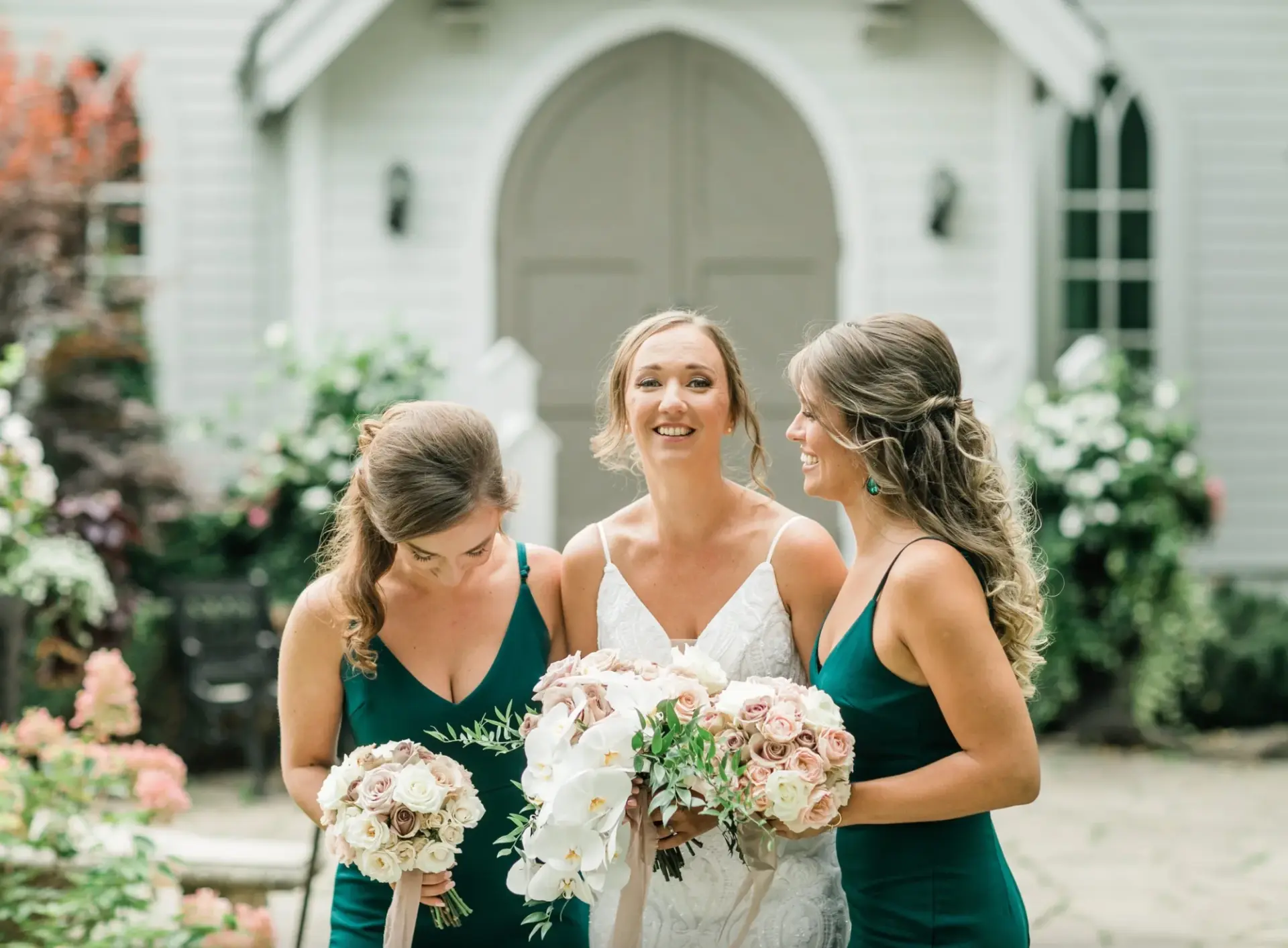 Kat laughing with her bridesmaids in green dresses, holding orchid and rose bouquets outside the chapel at The Doctor’s House