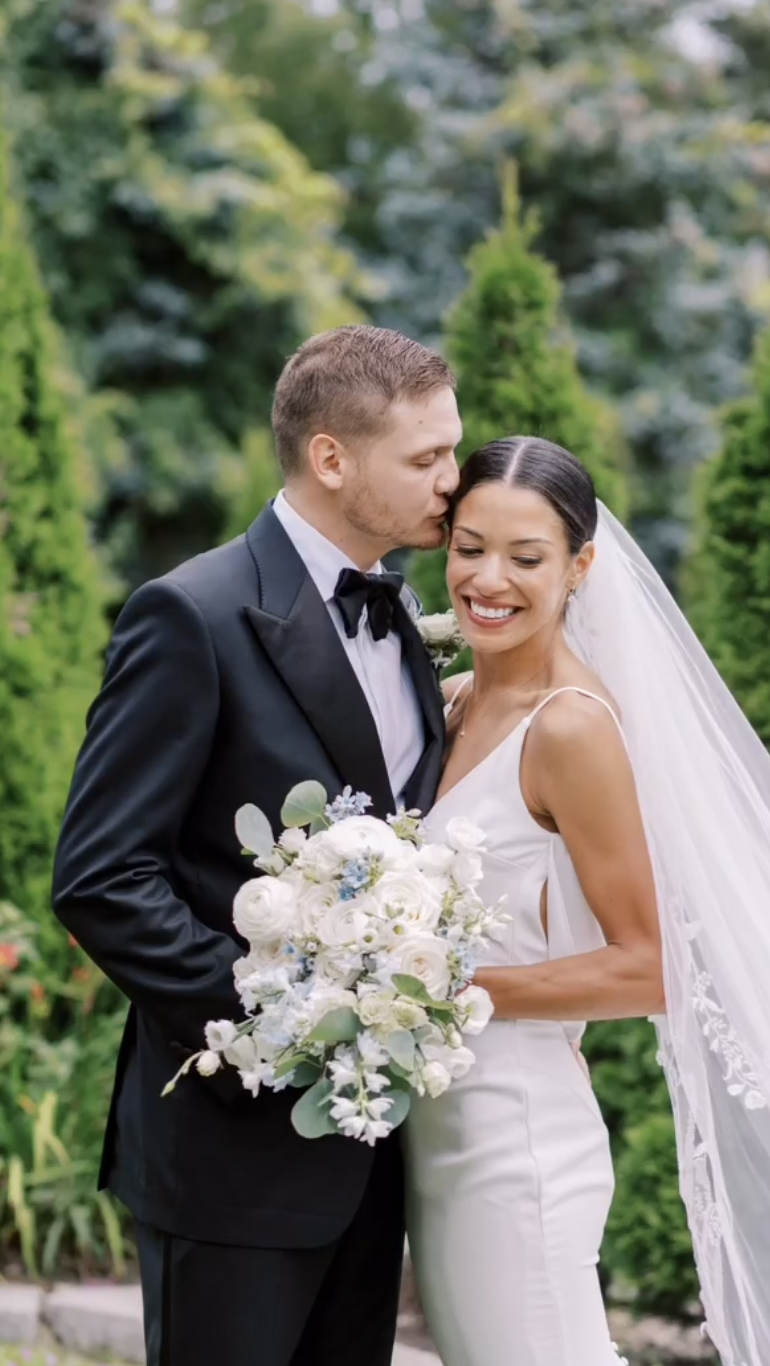 Groom kissing the bride’s forehead as she holds a white rose and blue delphinium bouquet