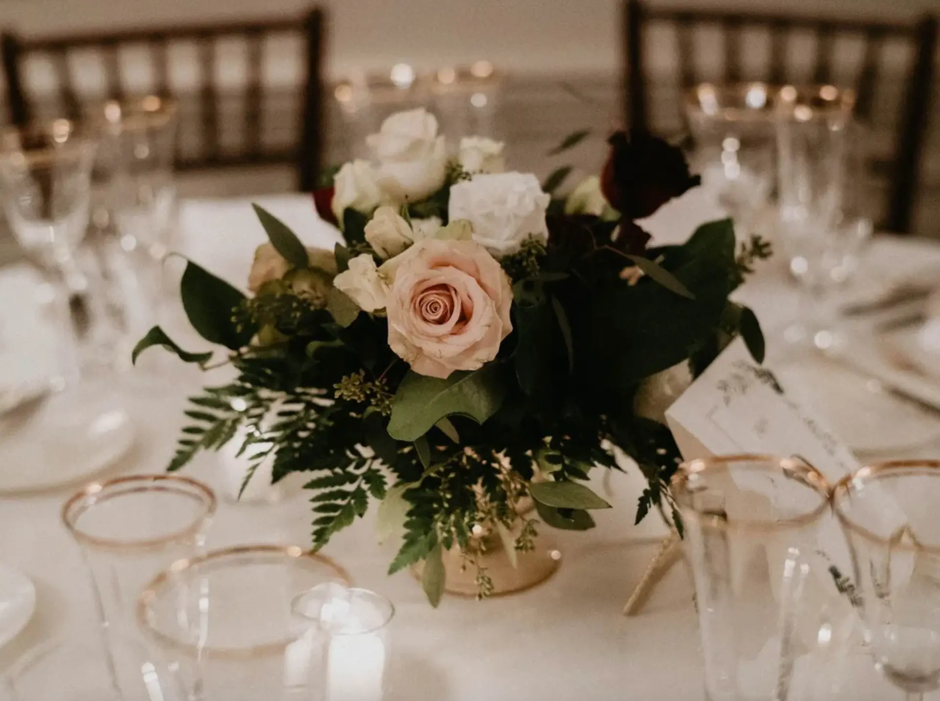 Low centrepiece of blush and ivory roses with ferns in a gold vessel among gold-rimmed glassware